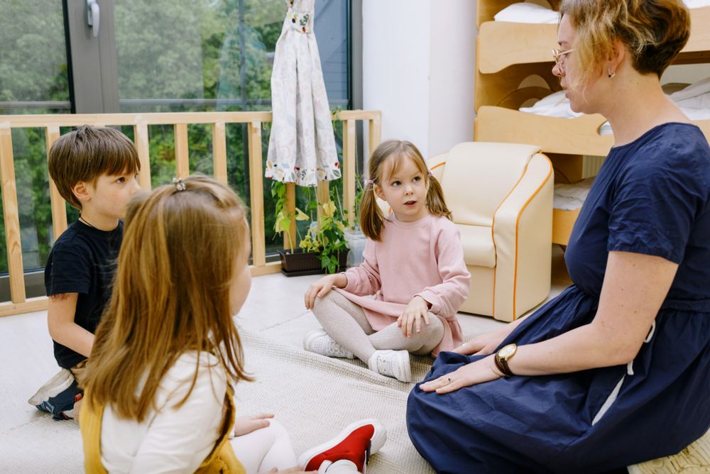 A teacher sitting on the floor talking with two young children in a bright room