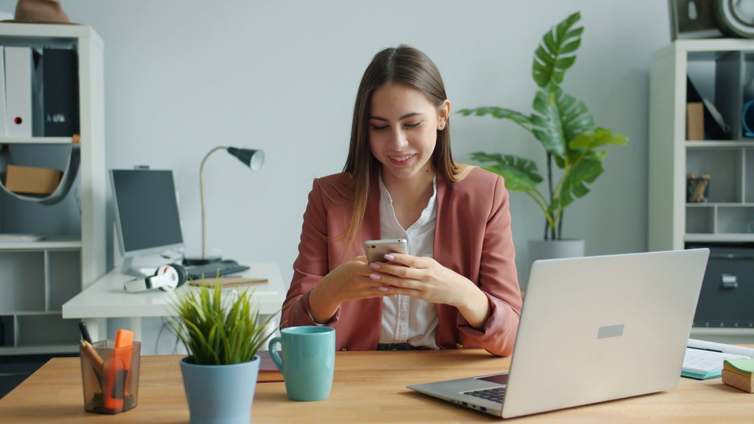 A woman at her desk with a laptop and phone, smiling