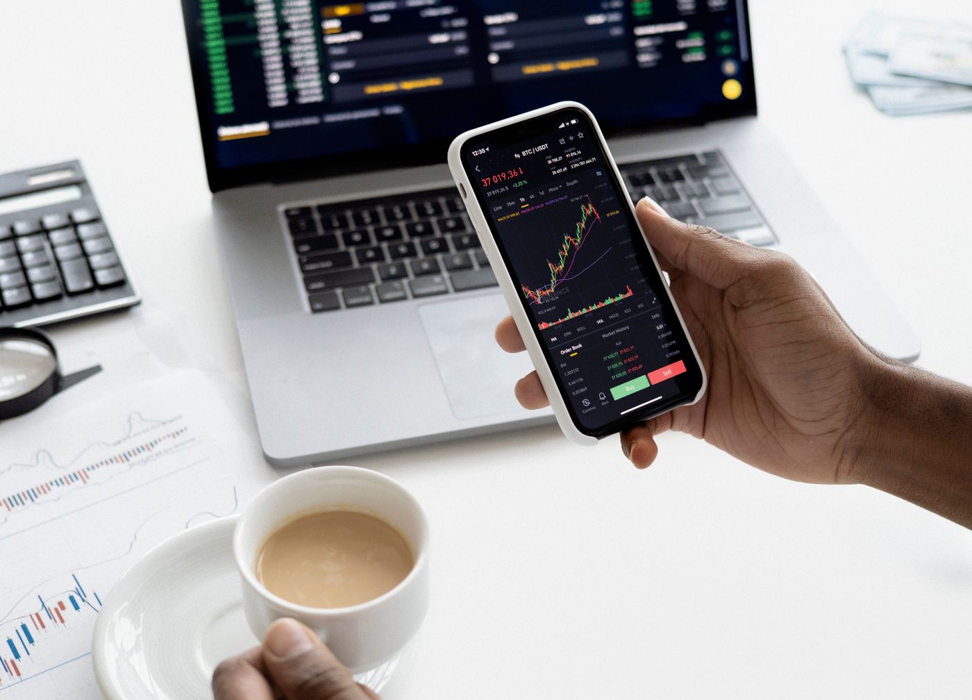 Hand holding a phone showing a growth chart, laptop with analytics behind it